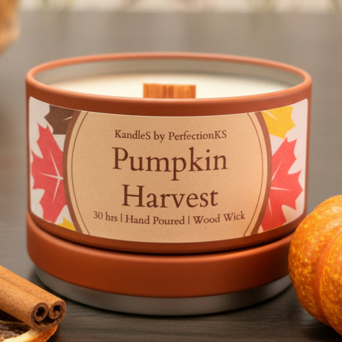 A candle tin labeled 'Pumpkin Harvest' by CandleS by PerfectionKS, with a terracotta-colored metal container, cream wax, and a decorative label featuring autumn leaves, set on a dark tabletop with a blurred floral background.