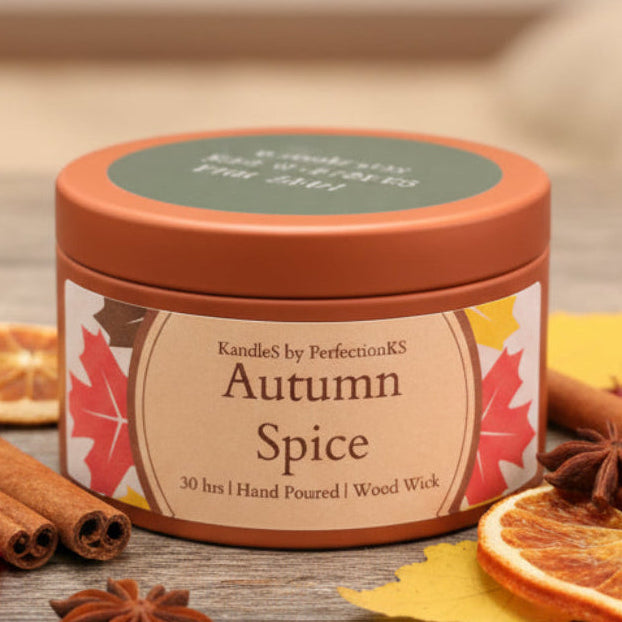 A round metal candle tin labeled 'Autumn Spice' by KandleS by PerfectionKS, sitting on a dark wooden surface with a blurred background.