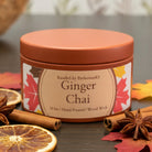 A round terracotta-colored candle tin labeled 'Saffron Spice' by Kandles by PerfectionKS, sitting on a dark wooden surface with a softly blurred background.