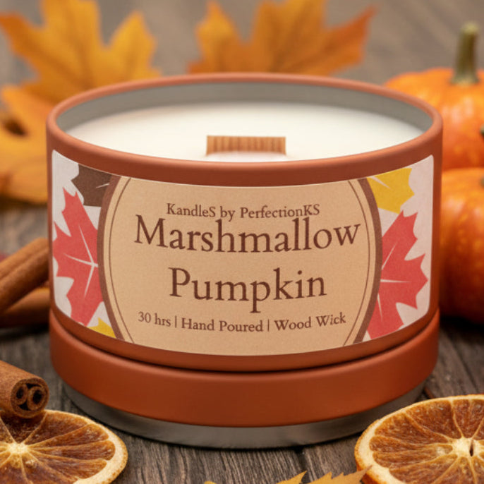A candle tin labeled 'Marshmallow Pumpkin' by Kandles by PerfectionKS, with a terracotta-colored lid and base, white wax, and fall leaf graphics on the label, set on a dark wooden surface.