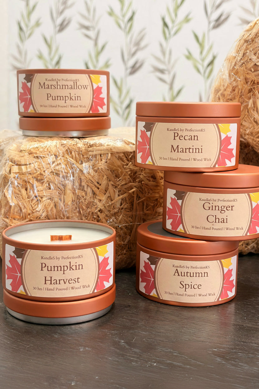 Five scented wood-wick candles in round copper-toned metal tins are arranged on a dark tabletop with decorative straw and a leafy wall backdrop. Visible labels include Pumpkin Marshmallow, Pecan Martini, Autumn Spice, Saftron Spice, and Pumpkin Harvest.