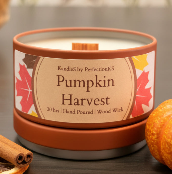 A candle tin labeled 'Pumpkin Harvest' by CandleS by PerfectionKS, with a terracotta-colored metal container, cream wax, and a decorative label featuring autumn leaves, set on a dark tabletop with a blurred floral background.
