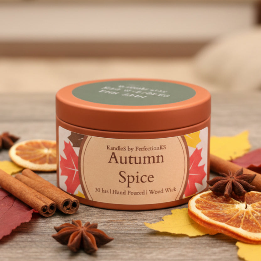 A round metal candle tin labeled 'Autumn Spice' by KandleS by PerfectionKS, sitting on a dark wooden surface with a blurred background.